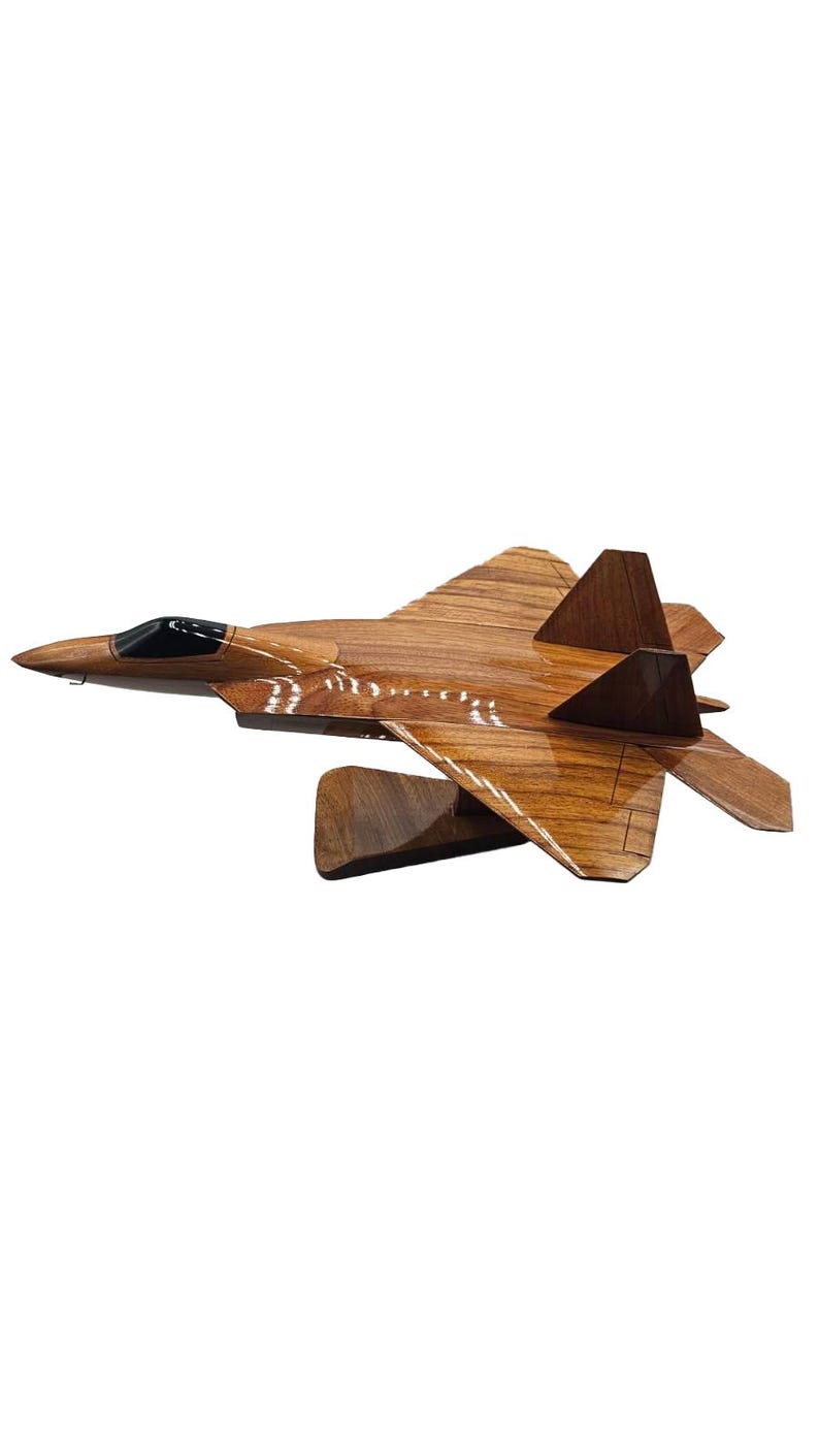 May include: A wooden model of a fighter jet, showcasing a detailed design with a brown finish. The jet features a sleek, aerodynamic shape, including wings, tail fins, and a cockpit. The model is mounted on a wooden stand.