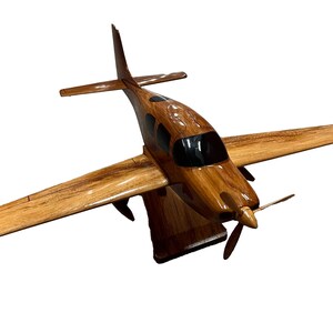 May include: A wooden model airplane with a brown and black paint scheme. The model is on a wooden stand and has a propeller.