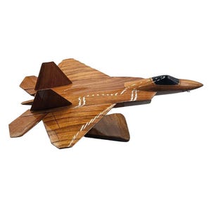 May include: A wooden model of a fighter jet, showcasing a detailed design with a brown finish and black cockpit. The wings and tail are angled, and the jet is mounted on a wooden stand. A decorative item.