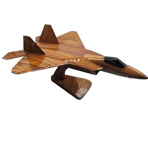 May include: A wooden model of a fighter jet, showcasing a detailed design with wings, tail fins, and a cockpit. The model is crafted from wood with a polished finish, mounted on a wooden stand.