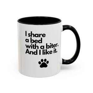 May include: White ceramic mug with a black interior and handle. The mug features the text "I share a bed with a biter. And I like it." in black, along with a black paw print.
