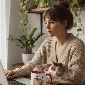 May include: A white ceramic mug with a red interior and handle, featuring a cartoon cat design with the text "Nap Zone, Recharge". The mug is on a wooden table, next to a laptop. A cat is resting on a person's lap.