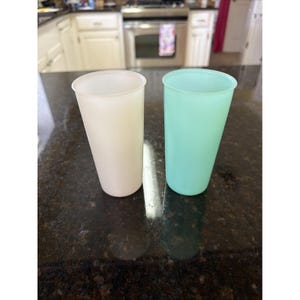 May include: Two tall, tapered plastic tumblers. One is a creamy off-white, the other a soft mint green. Both have a smooth, matte finish and a slightly flared rim. The tumblers are empty and sit on a dark, speckled countertop.