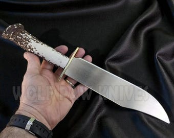 May include: A large hunting knife with a polished steel blade and a white handle with brown accents. The knife has a brass bolster and is displayed against a black background. The text "WOLF HOW KNIVES" is visible.
