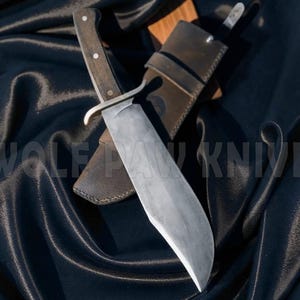 May include: A large, handmade knife with a dark wooden handle and a silver blade, resting on a black satin surface. The knife is accompanied by a brown leather sheath. The text "WOLF PAW KNIVES" is visible in the image.