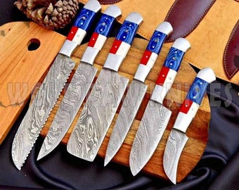 May include: Set of six Damascus steel knives with patterned blades. The handles feature a blue, white, and red design. The knives are arranged on a wooden cutting board, with a leather sheath visible in the background.