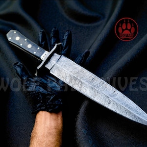 May include: A handcrafted knife with a patterned blade and a dark handle with silver accents. The knife is held against a black background. The text "WOLF PAW KNIVES" is visible.