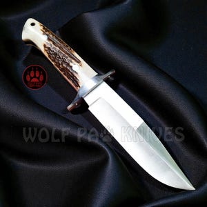 May include: A hunting knife with a polished steel blade and a handle made of bone and brown material. The knife has a silver bolster and a pointed tip. The text "WOLF PAW KNIVES" is visible.