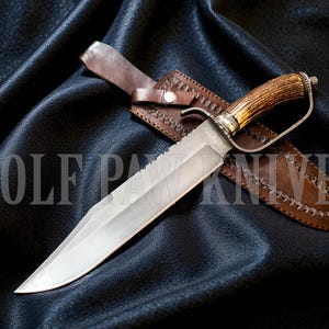 Può includere: Un grande coltello da caccia con una lama in acciaio lucido e un manico in materiale di corno. Il coltello ha una guardia ed è esposto con un fodero in pelle marrone. Il testo "WOLF PAW KNIVES" è visibile.
