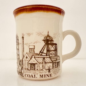 May include: Cream-colored ceramic mug with a brown rim and handle. The mug features a black and brown illustration of a coal mine with the words "COAL MINE" at the base. A vintage-style coffee mug.