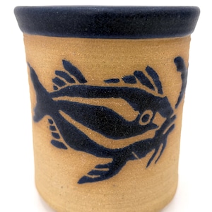 May include: A cylindrical ceramic mug with a tan body and a dark blue rim. A stylized dark blue fish design wraps around the mug. The mug is likely handmade and suitable for beverages.