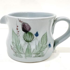 May include: A light blue ceramic creamer with a handle and spout. The creamer is decorated with a hand-painted floral design featuring a purple thistle, green leaves, and blue flowers. The creamer measures approximately 4 inches in height.