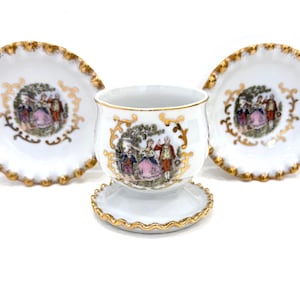 May include: A white porcelain egg cup with a matching saucer and two additional saucers, all featuring gold scalloped edges and a central colorful illustration of two figures in a garden setting surrounded by ornate gold scrollwork detailing.