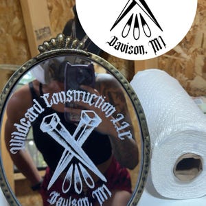 Może przedstawiać: Owalne lustro z ozdobną złotą ramą. Lustro odbija osobę trzymającą telefon i tekst "Wildcard Construction LLC Davison, MI" w okrągłym wzorze ze skrzyżowanymi gwoździami.