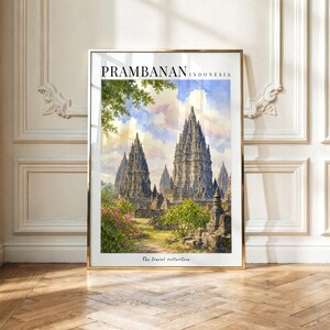 May include: A framed wall art print featuring a watercolor illustration of the Prambanan temple complex in Indonesia. The image displays tall stone spires under a blue sky with clouds, surrounded by green foliage and pink flowers. Text reads Prambanan Indonesia and The Travel collection.