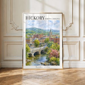 May include: A framed vertical art print featuring a watercolour painting of Hickory, North Carolina. The scene depicts a stone bridge over a river, lush green trees, and a town skyline with a clock tower under a blue sky. Text reads HICKORY NORTH CAROLINA.