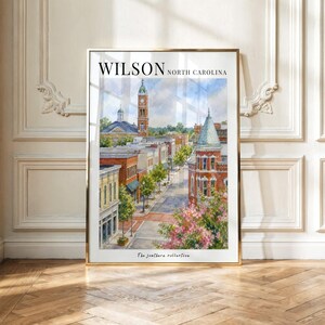 Puede incluir: Una impresión en acuarela enmarcada de una escena callejera en Wilson, Carolina del Norte. La obra muestra edificios de ladrillo, una torre de reloj y árboles verdes bajo un cielo azul. El texto dice WILSON NORTH CAROLINA y The southern collection.