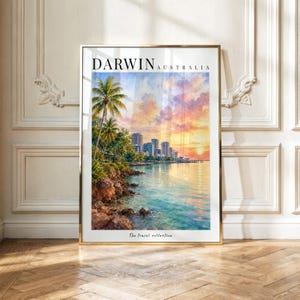 Op de afbeelding: Een ingelijste kunstdruk met een aquarel van de skyline van Darwin, Australië, bij zonsondergang. De afbeelding toont palmbomen, rotsachtige kliffen en stadsgebouwen die weerspiegeld worden in het water onder een levendige oranje en roze lucht.