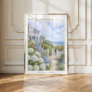 May include: A framed vertical art print titled THE HAMPTONS NEW YORK and The coastal collection. The image depicts a sandy path leading to the ocean, flanked by white picket fences, lush green foliage, and clusters of white and blue hydrangea flowers.