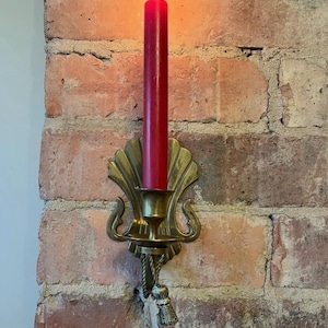 May include: A burning red candle in a brass wall sconce. The sconce features a decorative shell design and a tassel. The candle is long and thin, with a flame at the top. The sconce is fixed to a red brick wall.