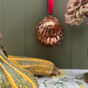 May include: A polished copper scallop shell mould featuring a reflective metallic surface and a scalloped edge. The mould is suspended by a vibrant red ribbon attached to a small metal loop at the top, creating a decorative hanging ornament.