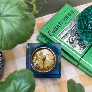 May include: A vintage clock with a gold-coloured face and black numerals is set in a square, transparent blue case. The clock sits on a yellow and white chequered surface, next to green books and a green glass ball.