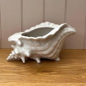 May include: A white ceramic seashell planter with a textured surface. The planter has a wide opening and is supported by small feet. The seashell design is detailed and realistic, ideal for coastal decor.