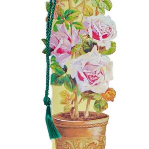 Pode incluir: Um marcador de página com uma ilustração vintage de um roseiral em vaso com flores rosa e brancas. O marcador de página tem uma borla verde.