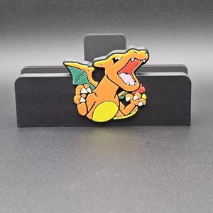 Puede incluir: Una colorida figura de Charizard, con detalles en naranja, amarillo, verde y rojo, está montada sobre un soporte de madera negro. La figura tiene la boca abierta y está en una pose dinámica. El soporte es rectangular con un soporte vertical.