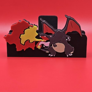 Peut inclure: Un organiseur de bureau noir avec une figurine Charizard en 3D. Le Charizard est marron avec des ailes rouges et un ventre crème, crachant des flammes jaunes et rouges. Le fond est rouge uni.