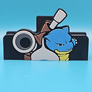 May include: A black, rectangular desk organizer featuring a cartoon character with blue, white, brown, and yellow accents. The design includes a circular element and a cylindrical shape, set against a light blue background.