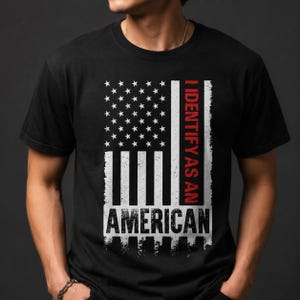 May include: Black t-shirt with a distressed American flag design. The flag features white stars and stripes, and the text "I IDENTIFY AS AN AMERICAN" in red and white. The shirt has a crew neck.