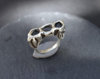 May include: A silver ring with a unique, textured design. The ring features three dark, possibly black, inset stones. The band has an organic, irregular shape, giving it a handcrafted appearance. The ring is displayed on a dark gray surface.