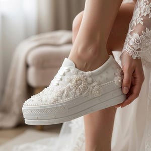 May include: White platform sneakers embellished with floral lace and pearl accents. The sneakers feature a thick sole and are designed with a classic lace-up style. The shoes are a bridal style, perfect for a wedding.