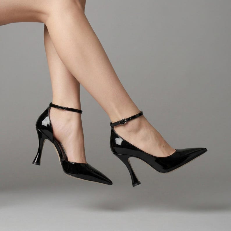 Peut inclure: Escarpins noirs en cuir verni avec bout pointu et bride de cheville. Les talons ont un design &eacute;vas&eacute; unique. Les chaussures sont brillantes et r&eacute;fl&eacute;chissent la lumi&egrave;re, sur fond gris.