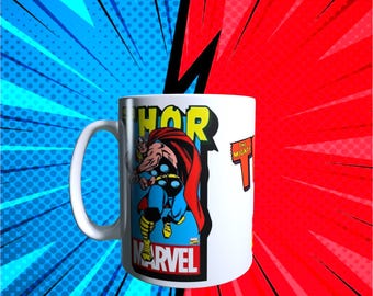 Könnte beinhalten: Weiße Keramik-Tasse mit einer Comic-Illustration von Thor, dem Wort "THOR" in Gelb und dem Marvel-Logo. Die Tasse hat einen weißen Henkel und die Worte "THE MIGHTY" auf der Seite. Der Hintergrund ist ein Comic-Design.