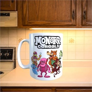 May include: A white ceramic mug featuring the black text Monster Cereals at the top and a colourful graphic below it. The illustration depicts a group of cartoon characters including a mummy, a blue ghost, a pink creature, a werewolf, and a vampire.
