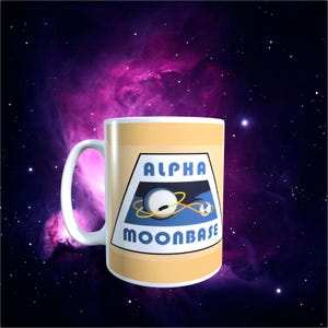 May include: A white ceramic mug with a light yellow band and a handle. The mug features a graphic with the words "ALPHA MOONBASE" and a design of a planet and a space station. The background is a nebula in space.