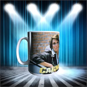 May include: White ceramic mug featuring a woman in a jacket, with the word "CAGNEY" in gold lettering. The background includes a brick wall and a blue and white striped design.