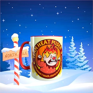 May include: A ceramic mug with a red handle and rim, featuring a cartoon Heat Miser character with the text "TEAM HEAT MISER" and "Whatever I touch, starts to melt in my clutch." The mug is set against a snowy winter scene.