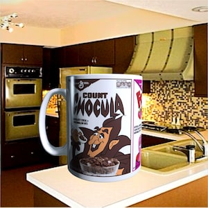 May include: A white ceramic mug featuring a graphic print of the Count Chocula cereal box, including the character illustration, the brand logo, and a bowl of chocolate cereal with marshmallow bits, set against a kitchen background.