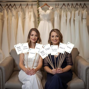 Może przedstawiać: Dwie kobiety w salonie sukien ślubnych trzymające tabliczki z napisami. Na tabliczkach widnieją napisy "Yes to the Dress", "Mein Favorit", "Hmm", "Uii!!", "Du siehst toll aus!", "Platz 2" i "Schön!". W tle wiszą suknie ślubne.