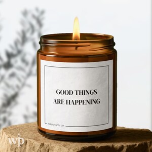 May include: A lit amber glass candle jar with a white label that reads "GOOD THINGS ARE HAPPENING." The candle sits on a stone surface. The flame is bright yellow and orange.