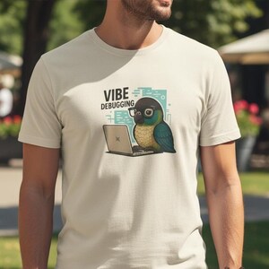 May include: Beige t-shirt with a cartoon parrot wearing glasses and working on a laptop. The text "VIBE DEBUGGING" is above the parrot. The parrot has blue, green, and yellow feathers.