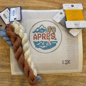 May include: A needlepoint canvas with the word "APRES" and a mountain scene, accompanied by skeins of blue, brown, and cream-colored thread, and a spool of yellow thread. The canvas is on a wooden surface.