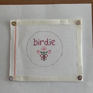 May include: A white needlepoint canvas with the word "birdie" stitched in pink letters inside a gray circle. A pink flower is stitched below the word. The canvas is secured to a white backing with four decorative buttons. A red and white stitched border is on the left side.