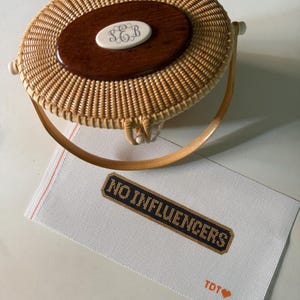 May include: A round woven rattan basket bag with a dark wood oval inlay featuring a white monogrammed plaque with the letters SEB. Below the bag lies a rectangular white needlepoint canvas with the black text NO INFLUENCERS and TDT with a heart.