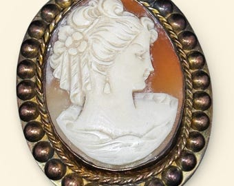 May include: Oval cameo brooch featuring a carved profile of a woman in white against an orange background. The cameo is set in a gold-toned frame with a beaded border and a silver-toned outer edge. The brooch is a vintage style.