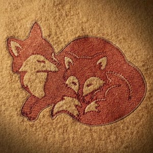 May include: A close-up of a tan textile with an embroidered design of two foxes. The foxes are in a reddish-orange color with cream-colored accents. The foxes are nestled together in a cozy pose.