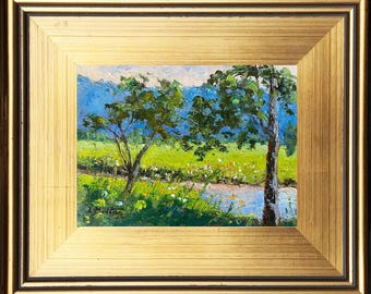 May include: A framed oil painting depicting a landscape scene with two trees in the foreground, a river, and a mountain range in the background. The trees are green and the river is blue. The mountains are blue and purple.